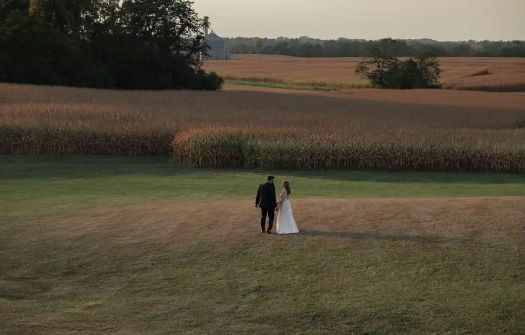 A couple in wedding attire holding hands in a serene rural landscape under the setting sun.